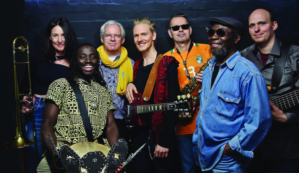 Die Band African Tam Tam. Sieben cool bis fröhliche Menschen vor einer blauen Wand